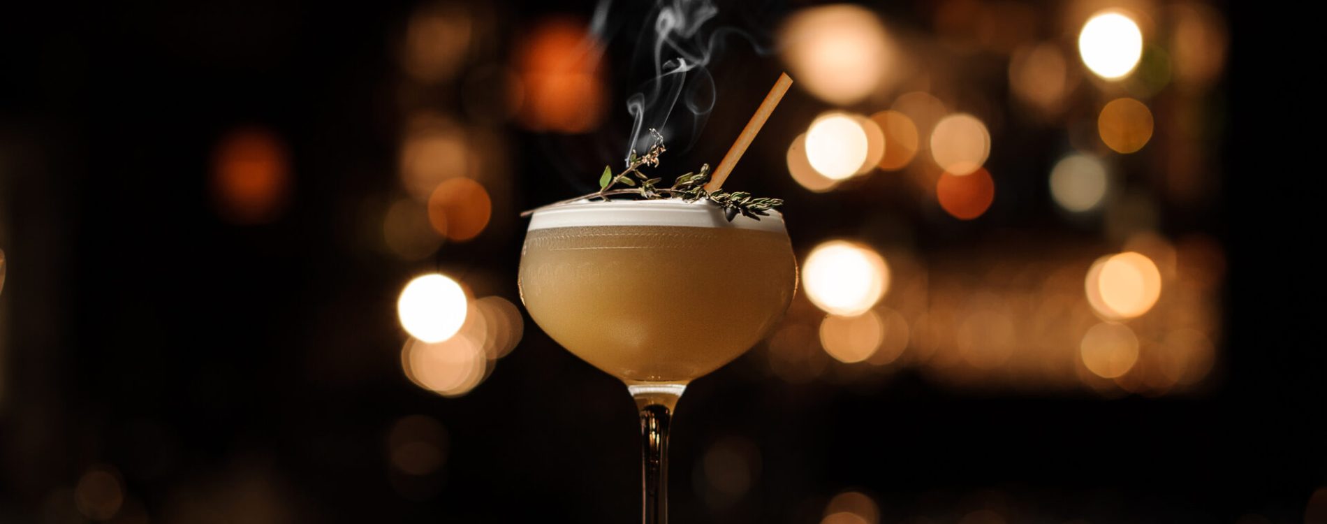 Delicious cocktail with a foam in the glass decorated with a smoked herb standing on the bar counter in the blurred background